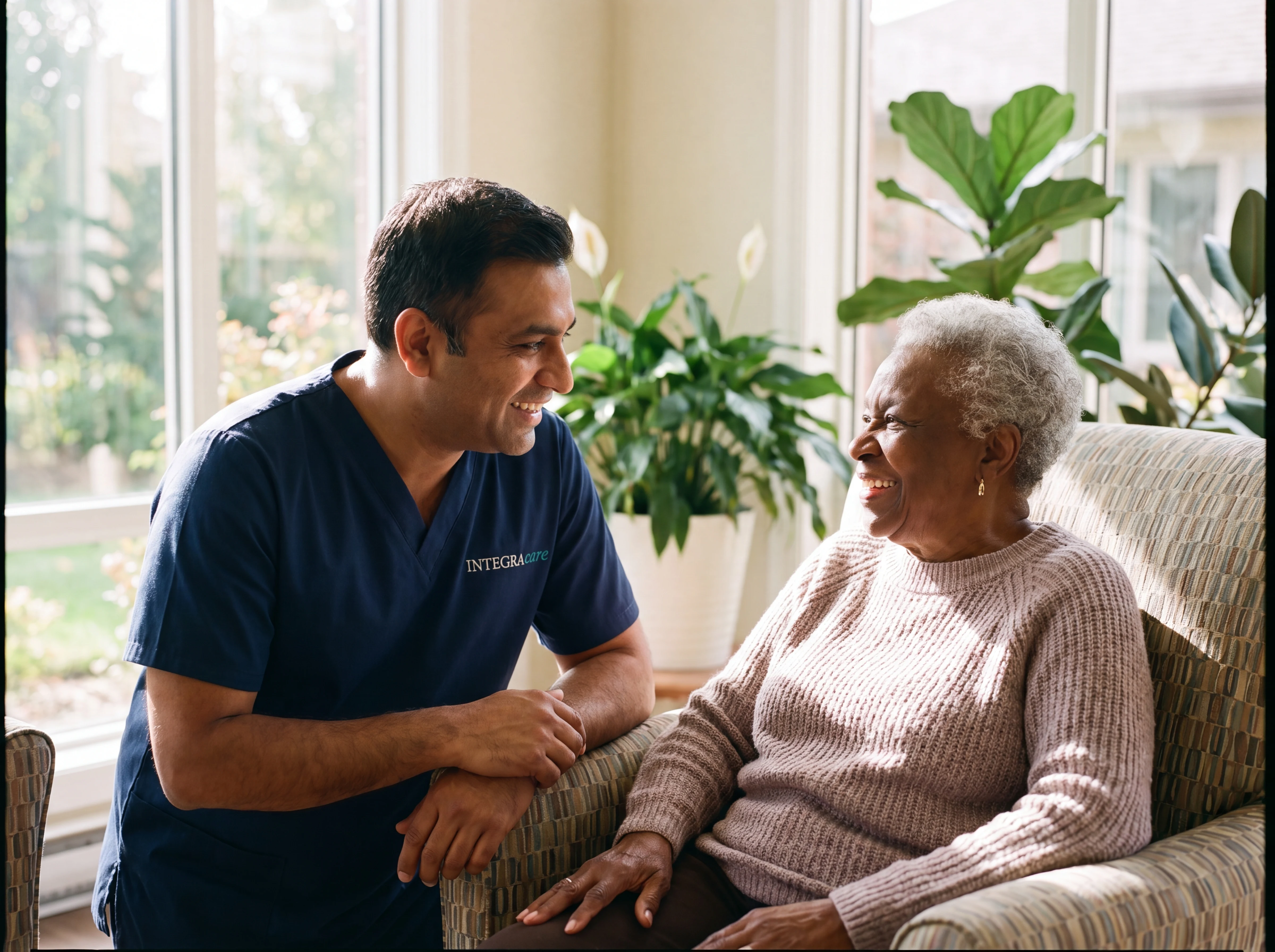 A South Asian male caregiver in relaxed conversation with an elderly Black woman in a bright retirement-residence lounge
